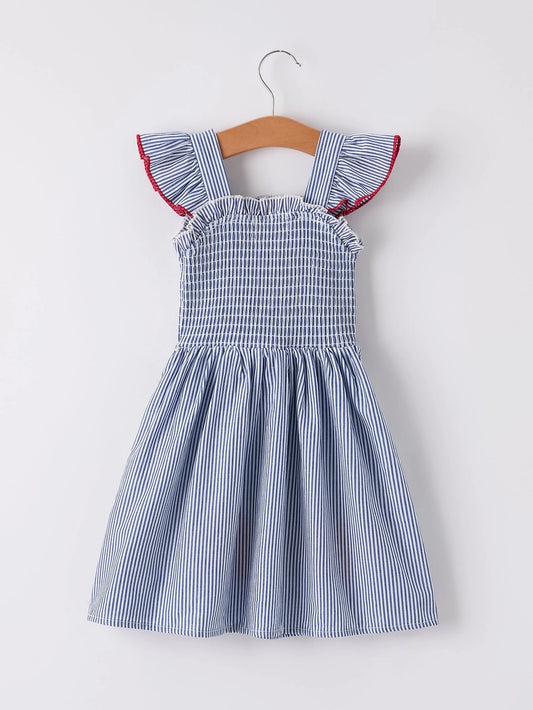 A blue and white striped sleeveless toddler dress with ruffled cap sleeves and red piping, hanging on a wooden hanger against a light gray background.