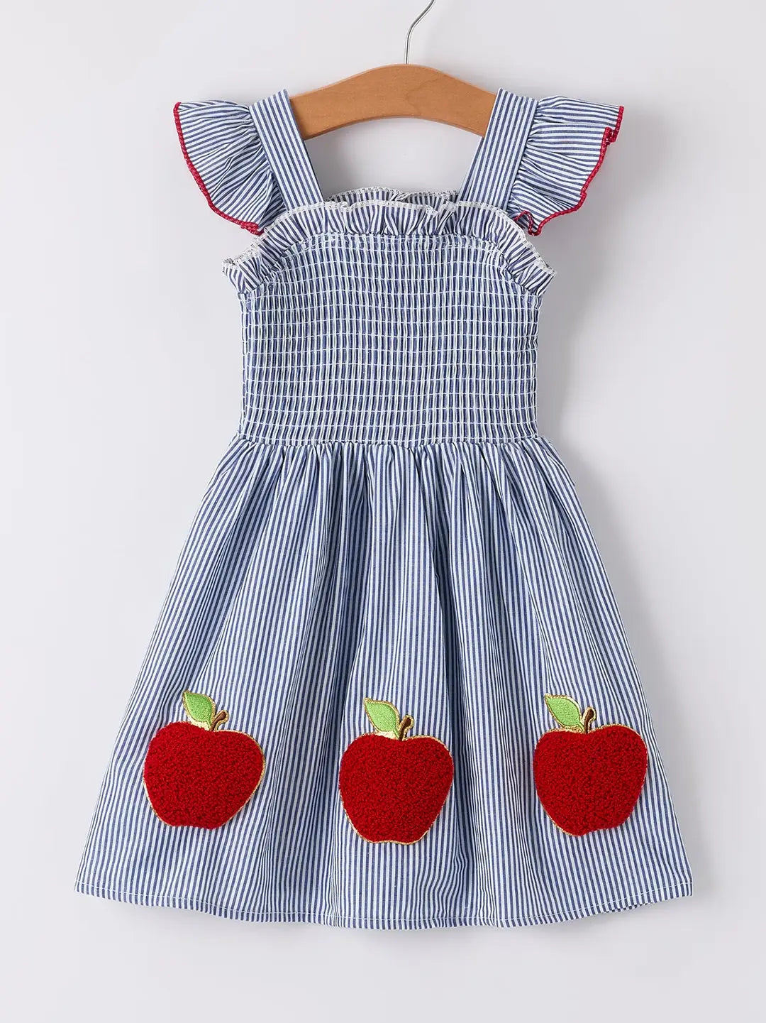A blue and white striped sleeveless dress for toddlers, featuring ruffled shoulder straps with red trim, a smocked bodice, and three red apple appliqués on the skirt. The dress is displayed on a wooden hanger against a plain white background.