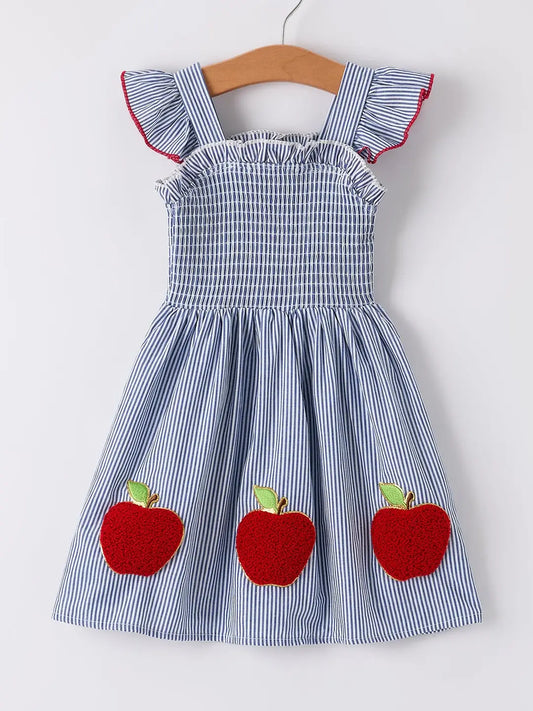 A blue and white striped sleeveless dress for toddlers, featuring ruffled shoulder straps with red trim, a smocked bodice, and three red apple appliqués on the skirt. The dress is displayed on a wooden hanger against a plain white background.